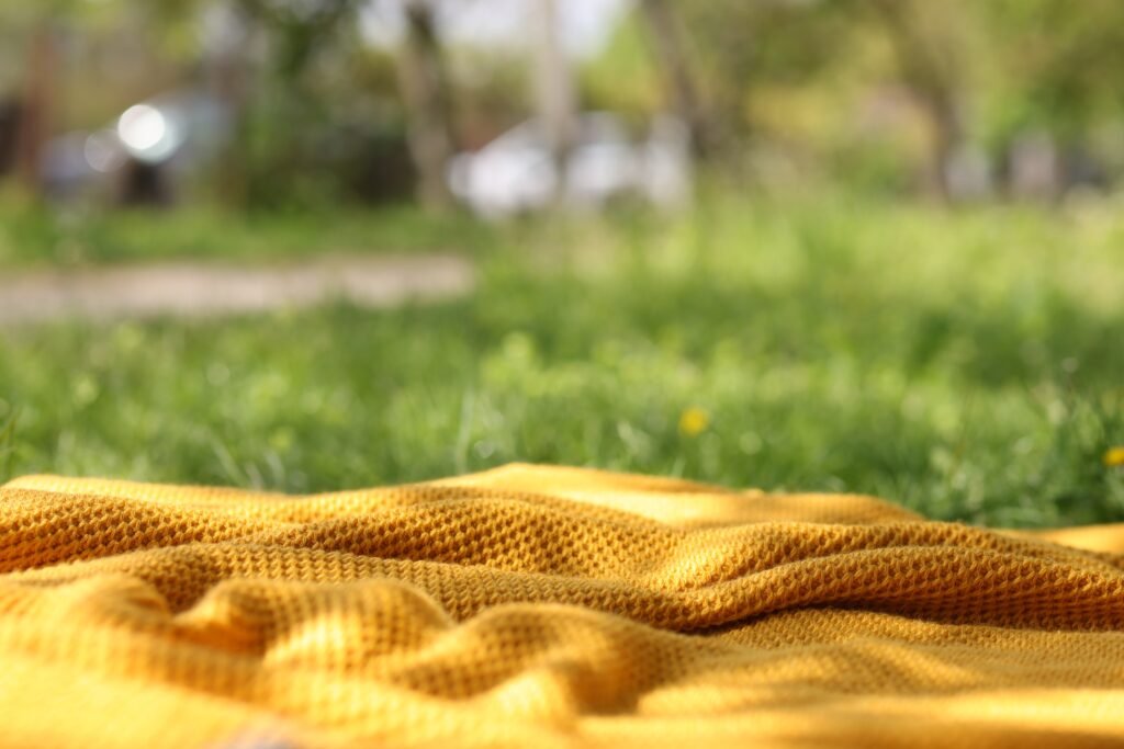 A soft-focus outdoor scene with a bright yellow rayon lawn fabric laid on green grass, sunlight filtering through trees in the background.