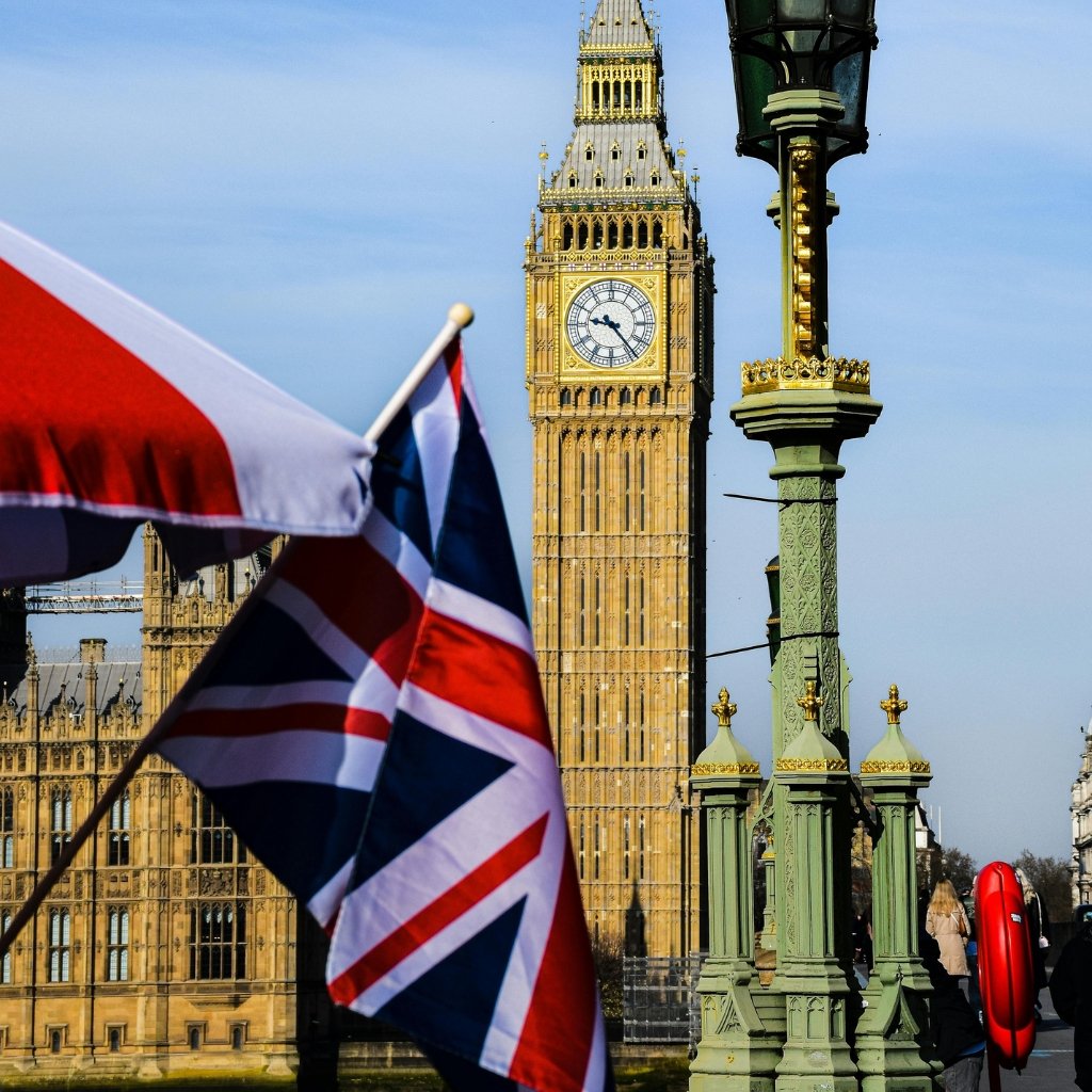 A close-up view of a Union Jack flag fluttering in the foreground with the Big Ben clock tower and the Houses of Parliament in the background.