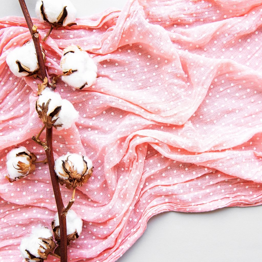 A pink fabric with white polka dots draped and folded, with a cotton branch featuring fluffy white cotton bolls resting on it.

