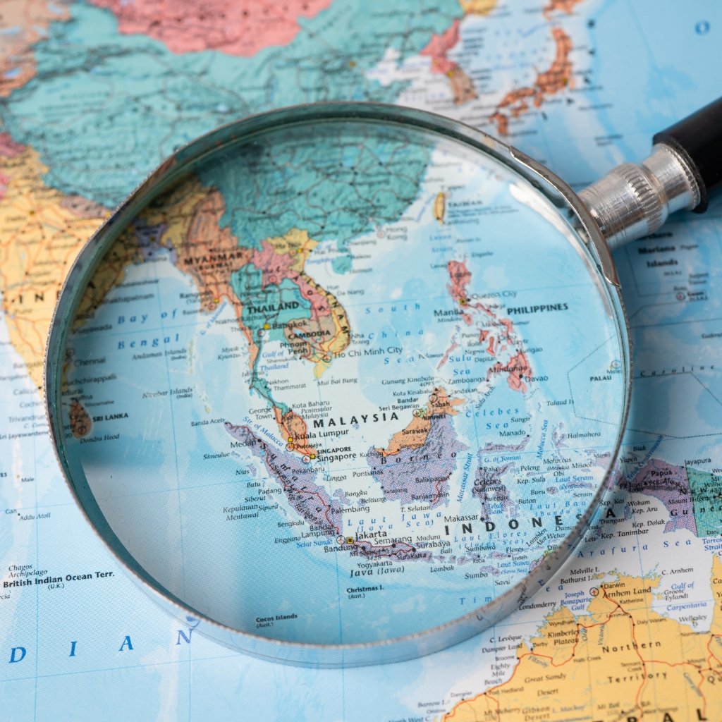 A close-up map of Southeast Asia viewed through a magnifying glass, focusing on countries like Malaysia, Thailand, Indonesia, and the Philippines.
