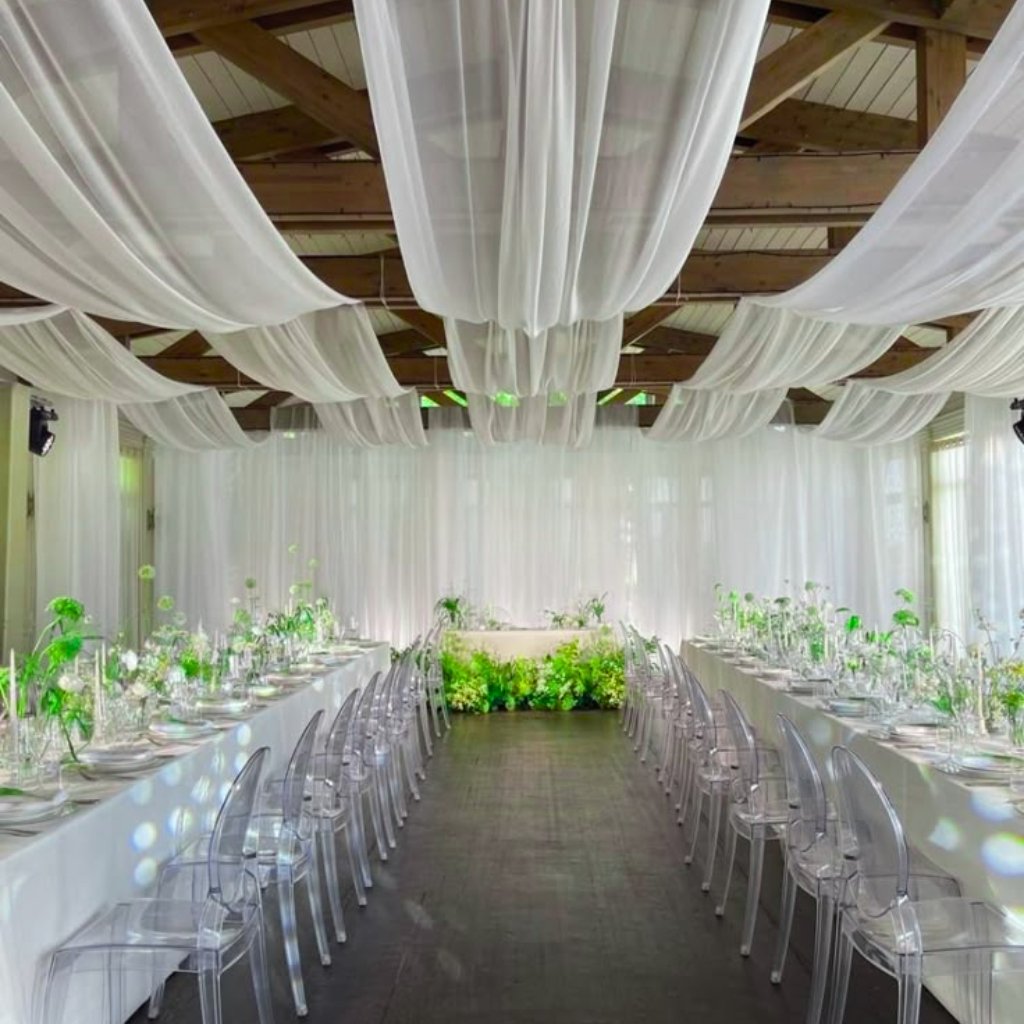 Elegant white event venue with long satin-organza draped ceiling swags and sheer panels. Two parallel rows of white-clothed tables with clear acrylic chairs, lush green floral centerpieces, and soft ambient lighting highlighting a bright, airy atmosphere