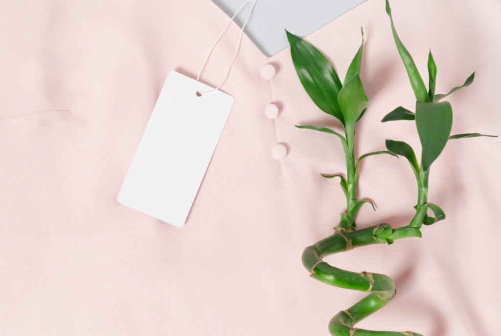 Close-up photo of a pink fabric background with a white blank tag attached by a string, and a green bamboo plant stem curling across the right side