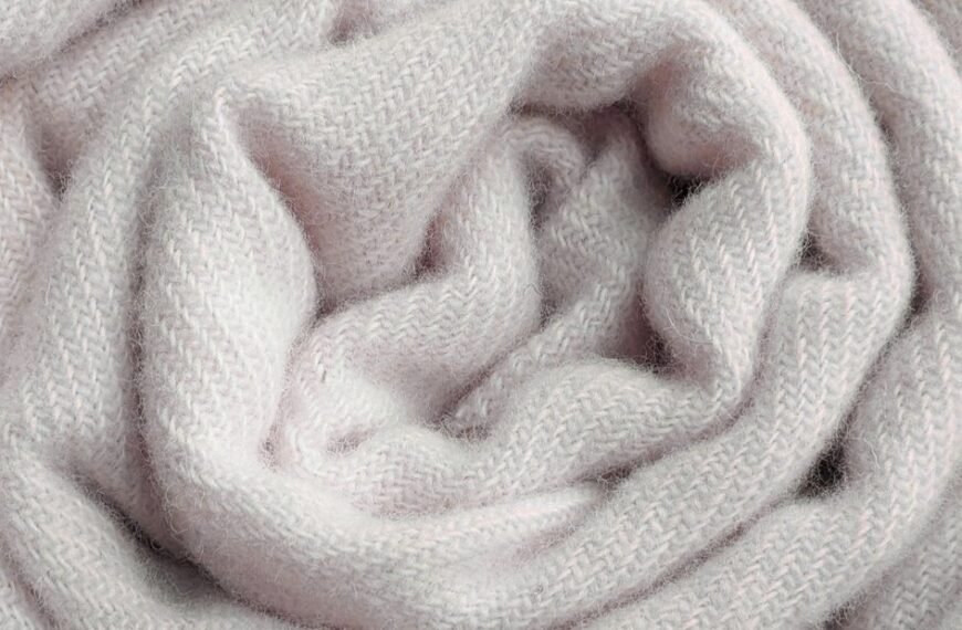 A close-up of a soft, pale pink knit scarf loosely twisted into folds, showing its textured woolen fabric.