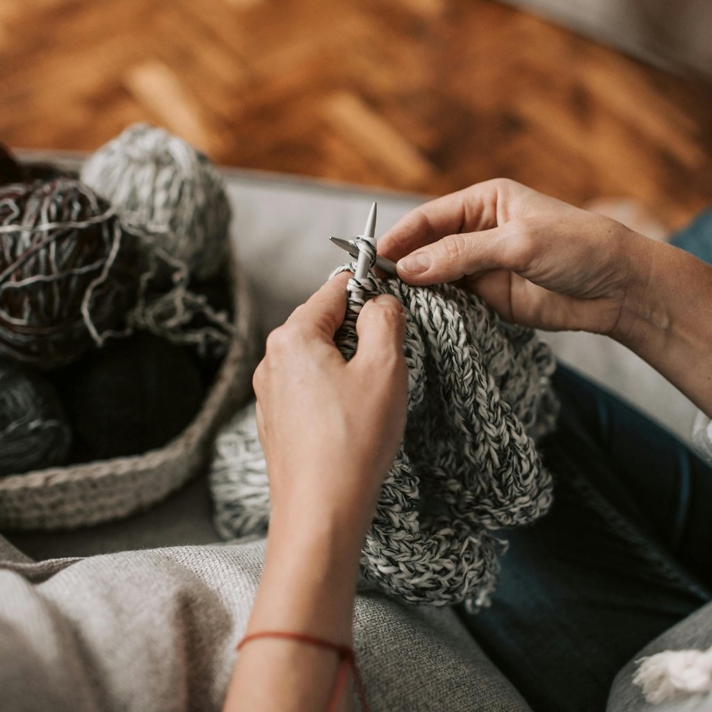 A close-up of hands knitting thick gray and white yarn on wooden knitting needles.
