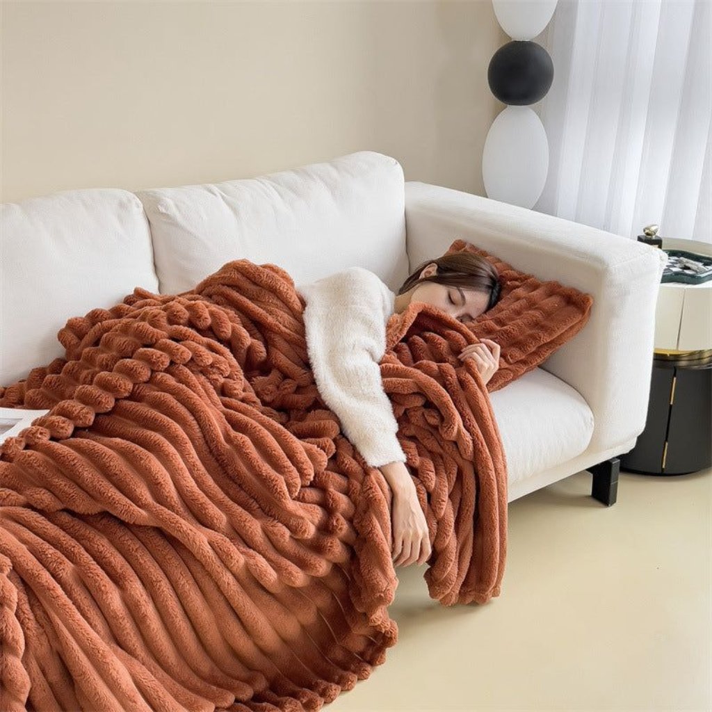 A person is curled up sleeping on a white sofa, covered with a chunky rust-colored blanket. The scene includes a stack of white and black balloons-style lamp in the background and a small side table.