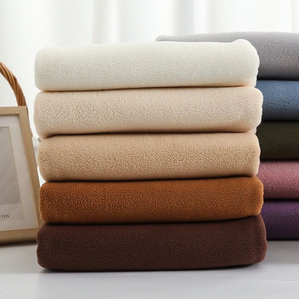 Stack of folded fuzzy blankets in neutral and warm tones (white, beige, light brown, tan, chocolate) on a white surface.