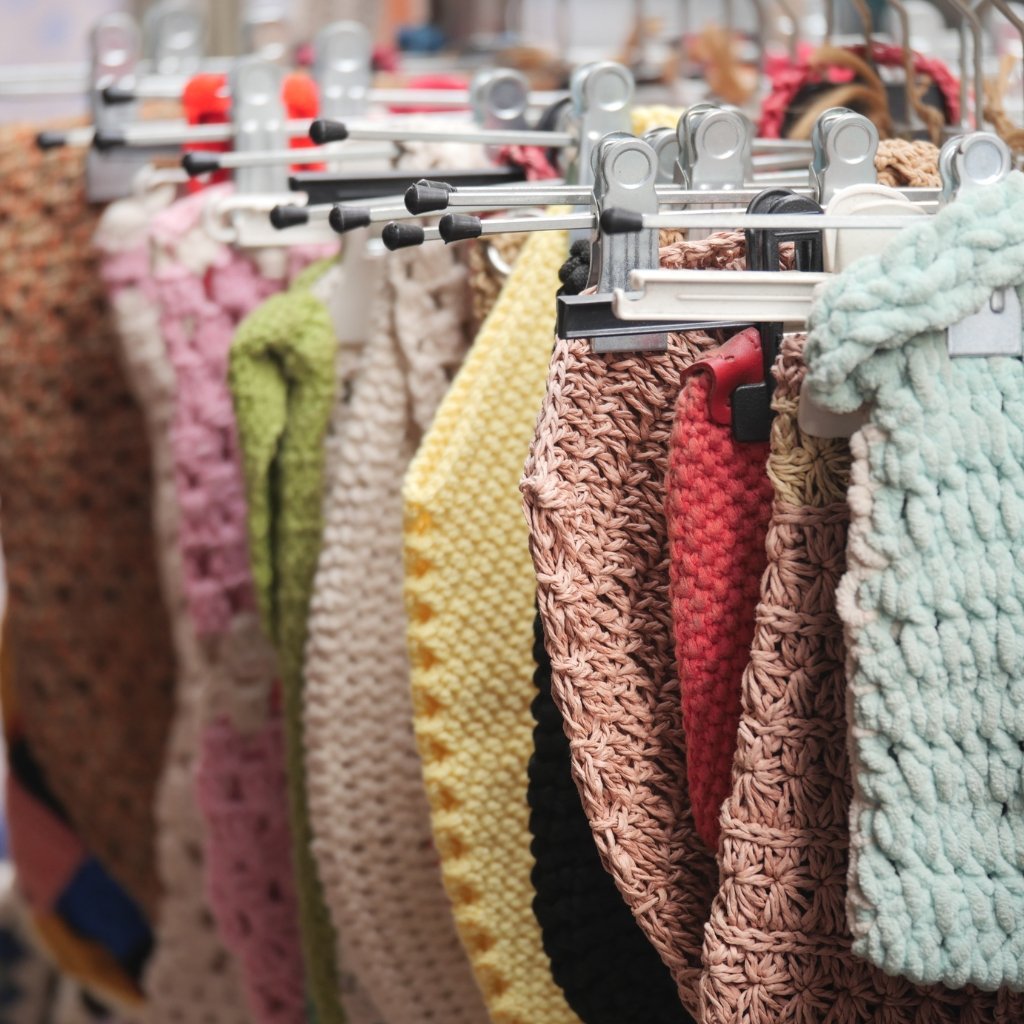 A row of chunky knitted scarves or blankets in pastel and earthy tones hanging on a rack with metal clips in a craft or yarn shop.