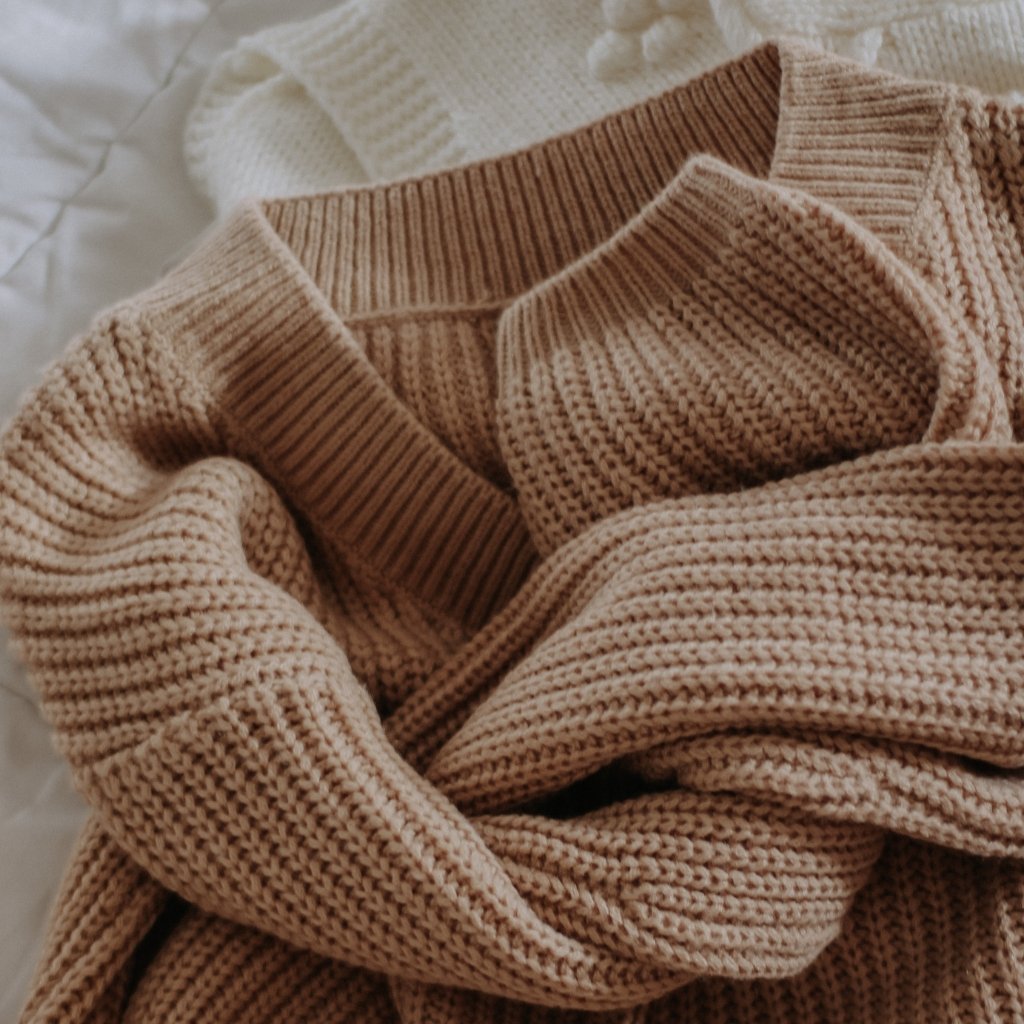A close-up of soft, chunky knit sweaters in warm beige and cream tones, folded and layered.