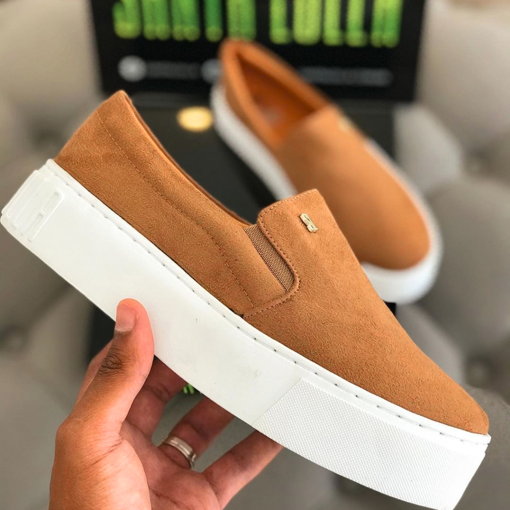 A tan suede slip-on sneaker with a thick white platform sole and elastic side inset, being held in a hand.