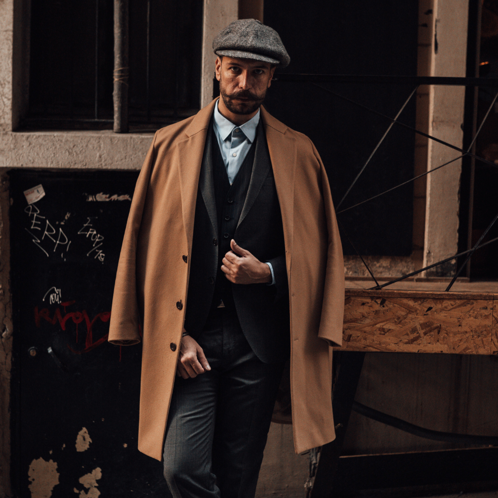 A man wearing a camel-colored overcoat draped over his shoulders, paired with a grey flat cap and a dark three-piece suit, standing in an urban alley.
