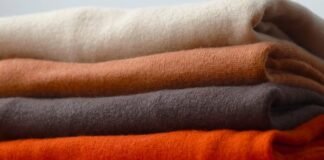 The Best Types of Wool for Clothing & Home Textile Explained A neatly stacked pile of folded sweaters in warm, earthy colors: cream, orange, olive, dark gray, and bright orange at the bottom. Best Types of Wool