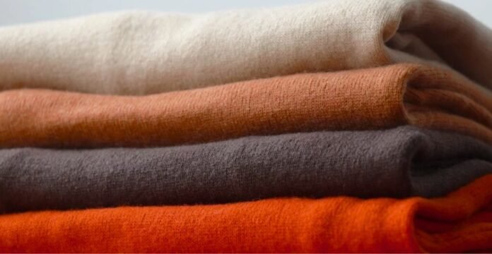 A neatly stacked pile of folded sweaters in warm, earthy colors: cream, orange, olive, dark gray, and bright orange at the bottom. Best Types of Wool