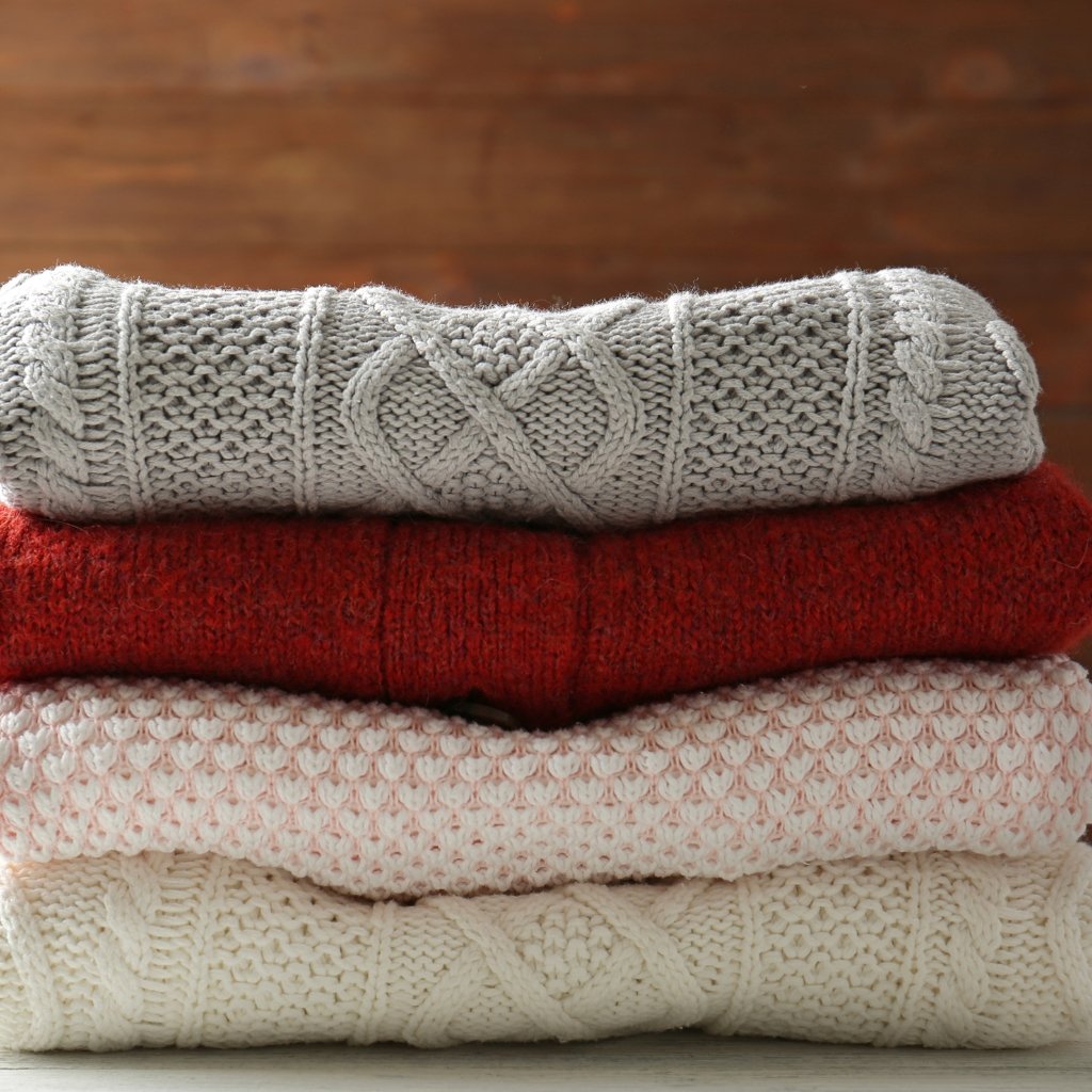 A stack of four knitted sweaters in varying colors (gray, red, light pink, cream) folded neatly on a wooden surface. Better Moisture Control