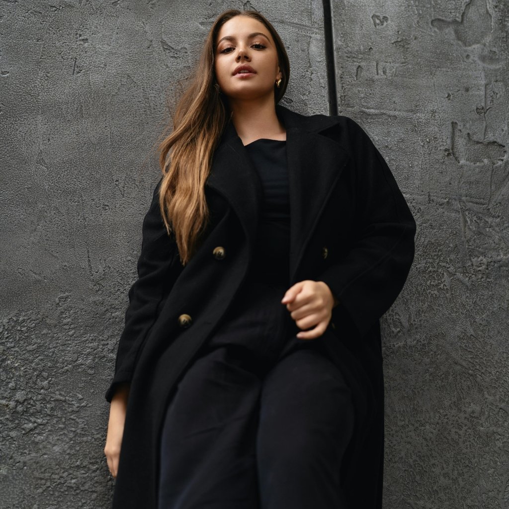 A young woman with long, light brown hair posing against a textured gray wall. She wears an oversized black coat with gold buttons and a black outfit underneath, looking confidently at the camera.