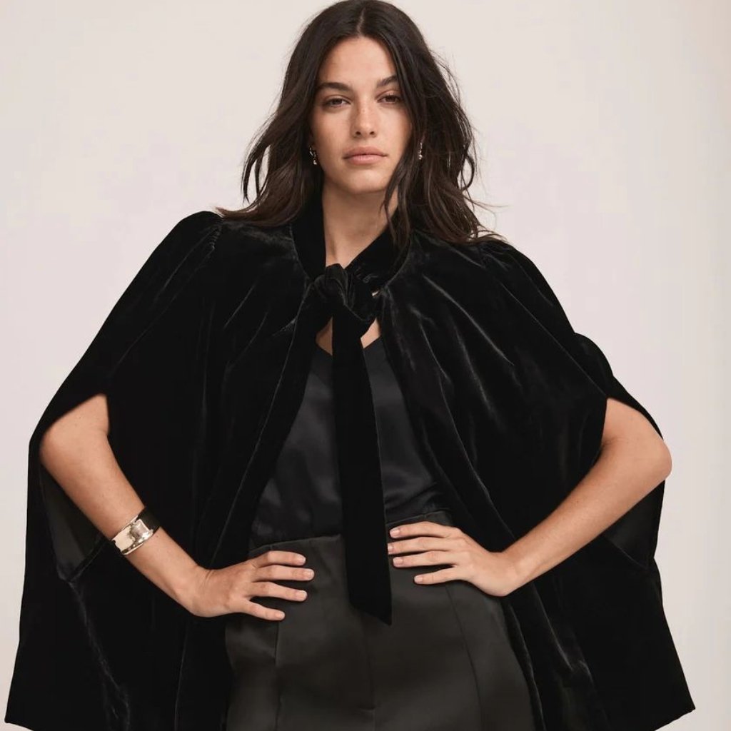 A stylish woman stands confidently in a luxurious black velvet cape with a tied bow at the neck, paired with a sleek satin top and a dark skirt.