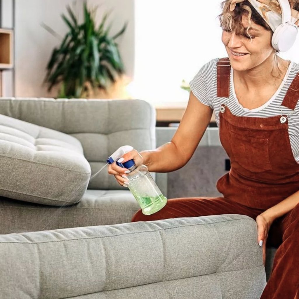 A person wearing red overalls and a striped shirt sprays a green cleaning solution onto a light gray sofa cushion while sitting on the arm of the couch and wearing white over-ear headphones.