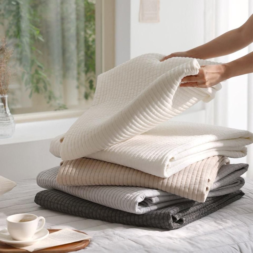 A stack of neatly folded ribbed blankets in neutral colors (cream, beige, taupe, light gray, charcoal) on a bed, with someone folding the top blanket. A cup of coffee and a small book sit nearby near a bright window.