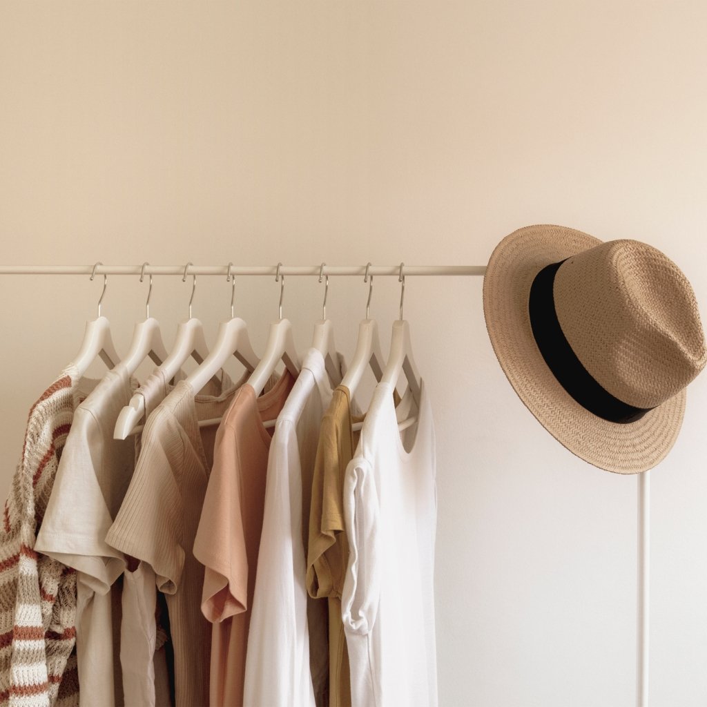 A minimalist clothing rack with a row of light-colored shirts and garments hanging on white hangers. A straw hat with a black band rests on the right end of the rack. The background is a plain, light-colored wall.