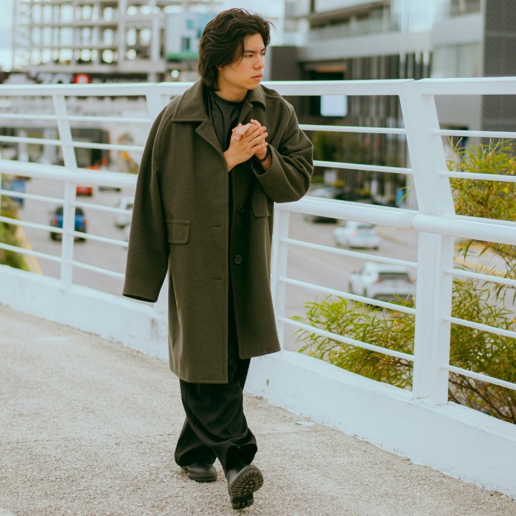 : A young man walks along a city pedestrian     path, wearing a long dark green coat over black clothing. He clasps his hands near his chest and looks contemplatively to the side, with a white railing and urban buildings in the background. Types of overcoat.