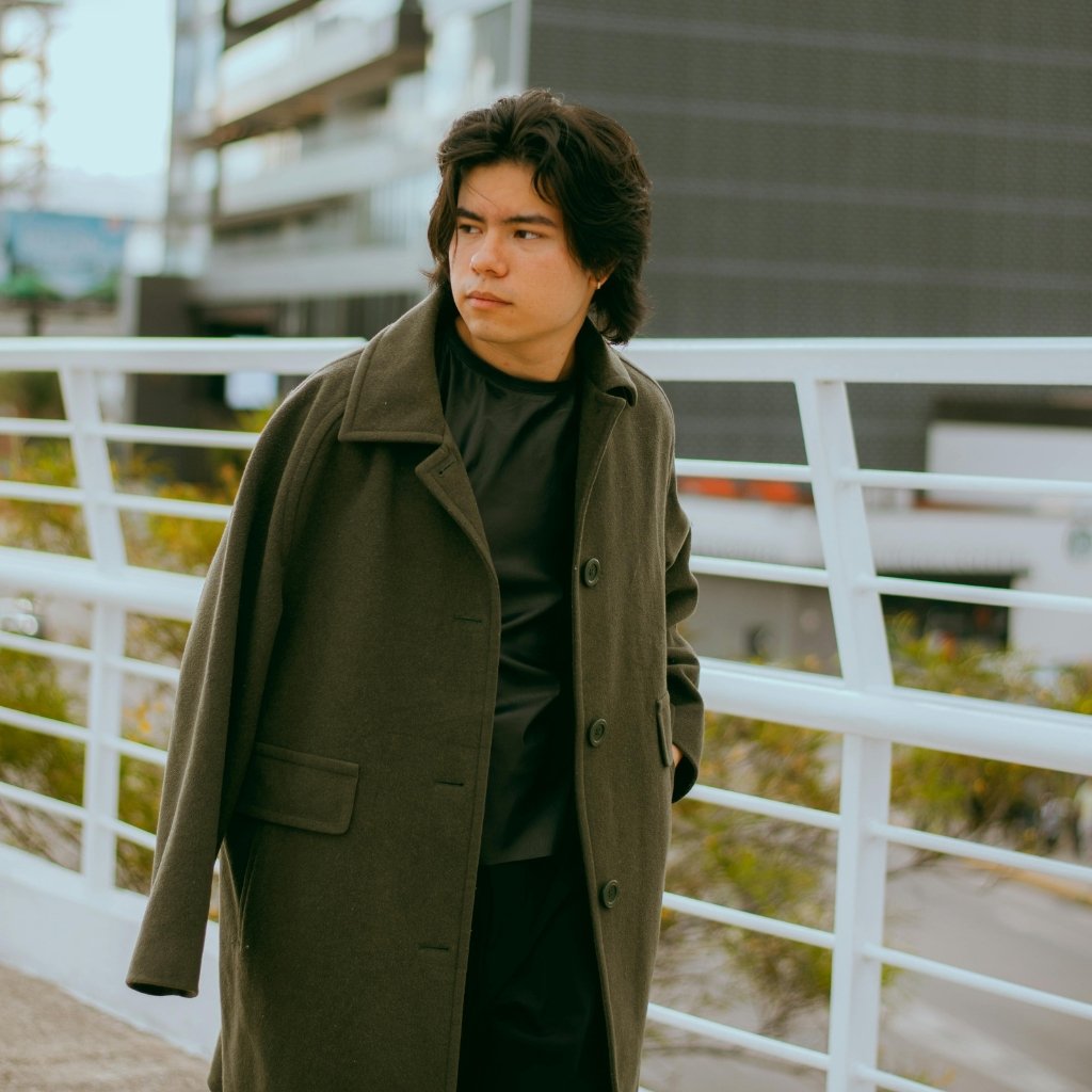 A man in a dark olive coat standing outdoors near a white railing, looking off to the side with a thoughtful expression.