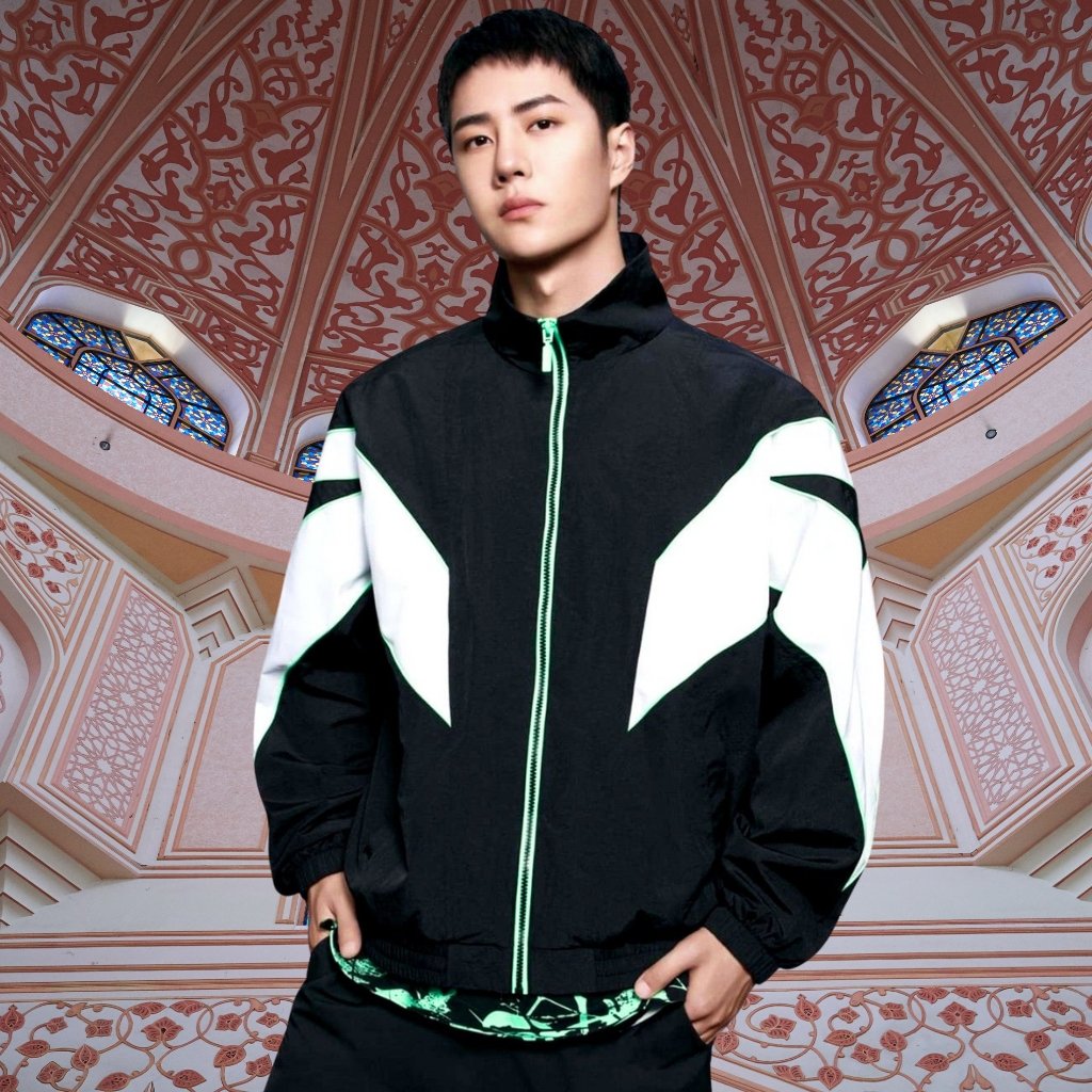 A young man wearing a black and white color-block windbreaker with bright green zippers stands in an ornately patterned, pink-toned architectural interior. He has short dark hair and a neutral expression, with hands in the jacket pockets.