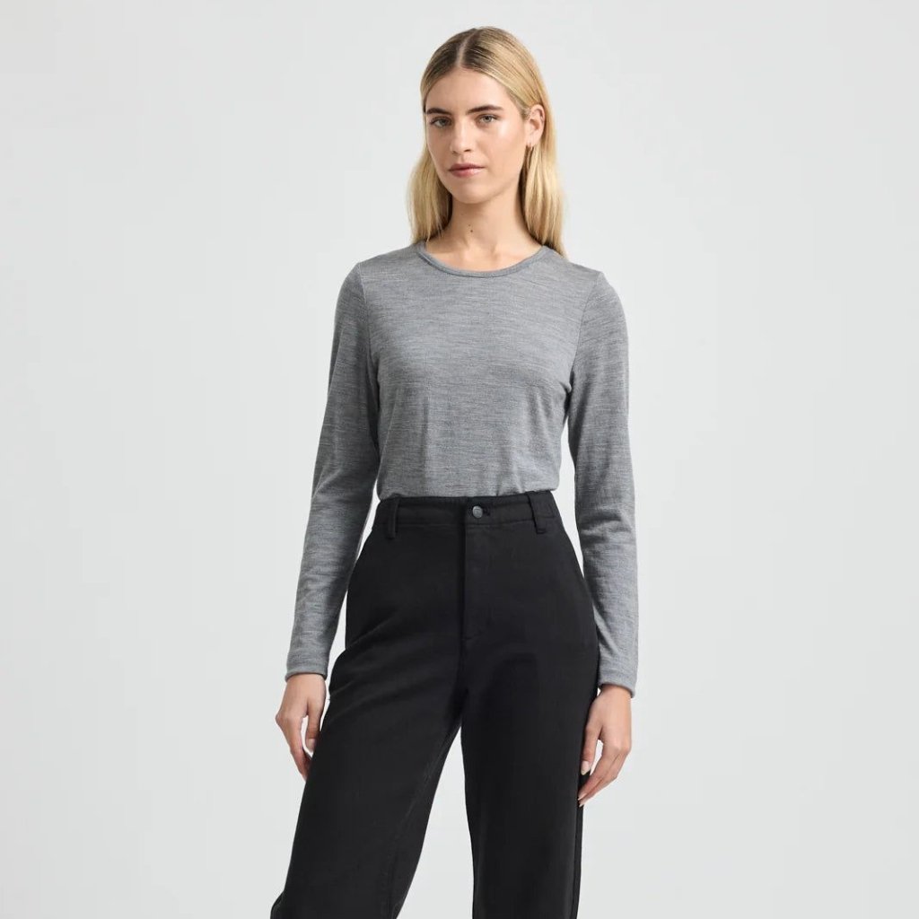 A young woman stands against a light gray backdrop, wearing a light gray long-sleeve top tucked into high-waisted black pants.