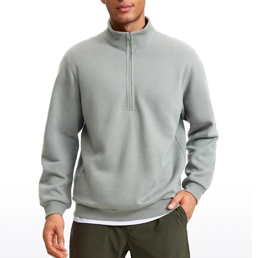A man wearing a light gray quarter-zip pullover sweatshirt with a high collar, standing with one hand in his pocket. He has a white layer peeking out from under the hem and is wearing olive-green pants.