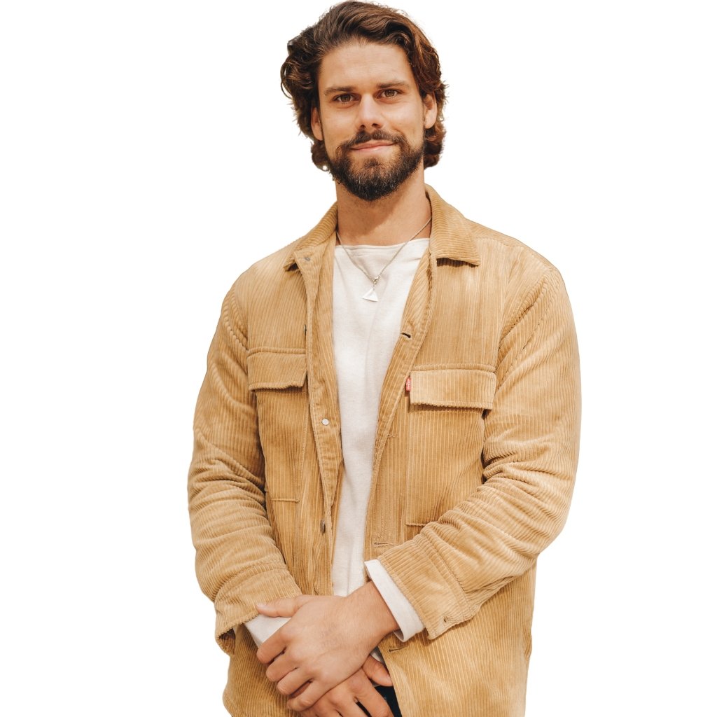 A man with a trimmed beard and wavy brown hair stands facing the camera, wearing a light beige corduroy jacket over a white tee. He has a relaxed smile and his hands clasped in front.