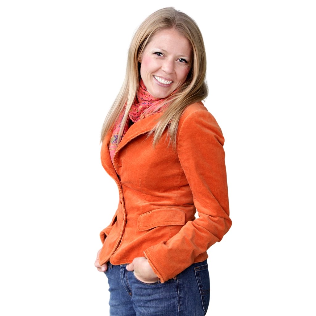 A smiling woman with long blonde hair wearing an orange blazer and a pink scarf, standing with hands in pockets against a white background.
