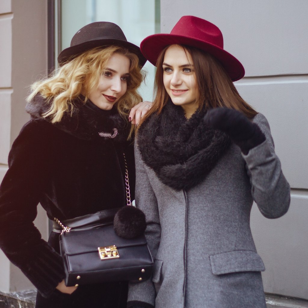 Two stylish women wearing winter coats and hats pose together on a city street. One woman in a black fur coat with a black hat holds a black handbag with a chain; the other wears a gray coat with a black scarf and a burgundy hat.The types of overcoat.