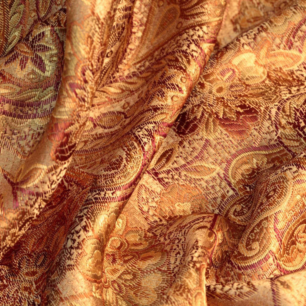 Close-up of luxurious brocade fabric with intricate floral and scroll patterns in warm gold, pink, and bronze tones.