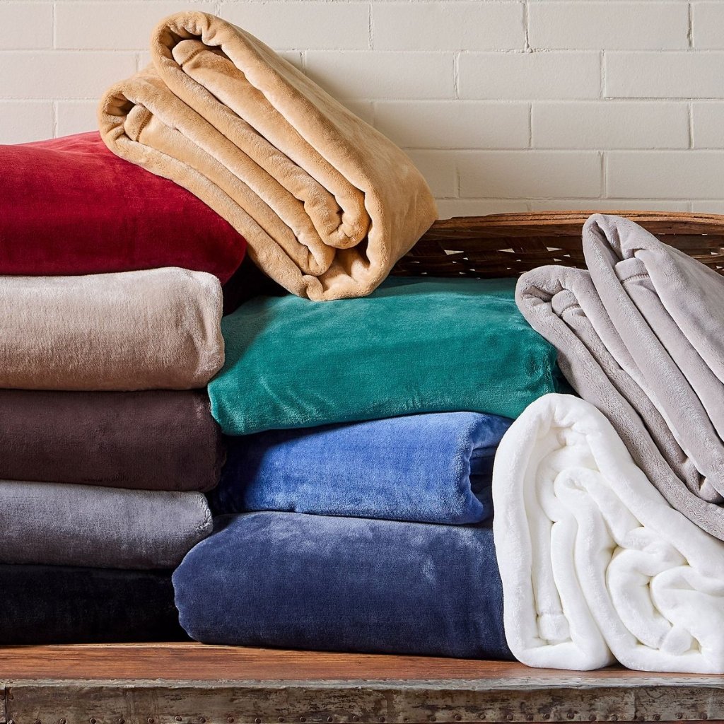 A stack of folded blankets in various colors (beige, red, tan, teal, blue, gray, and white) sits on a wooden surface against a white brick wall; some blankets are rolled at the top and one white fleece is draped on the right.

