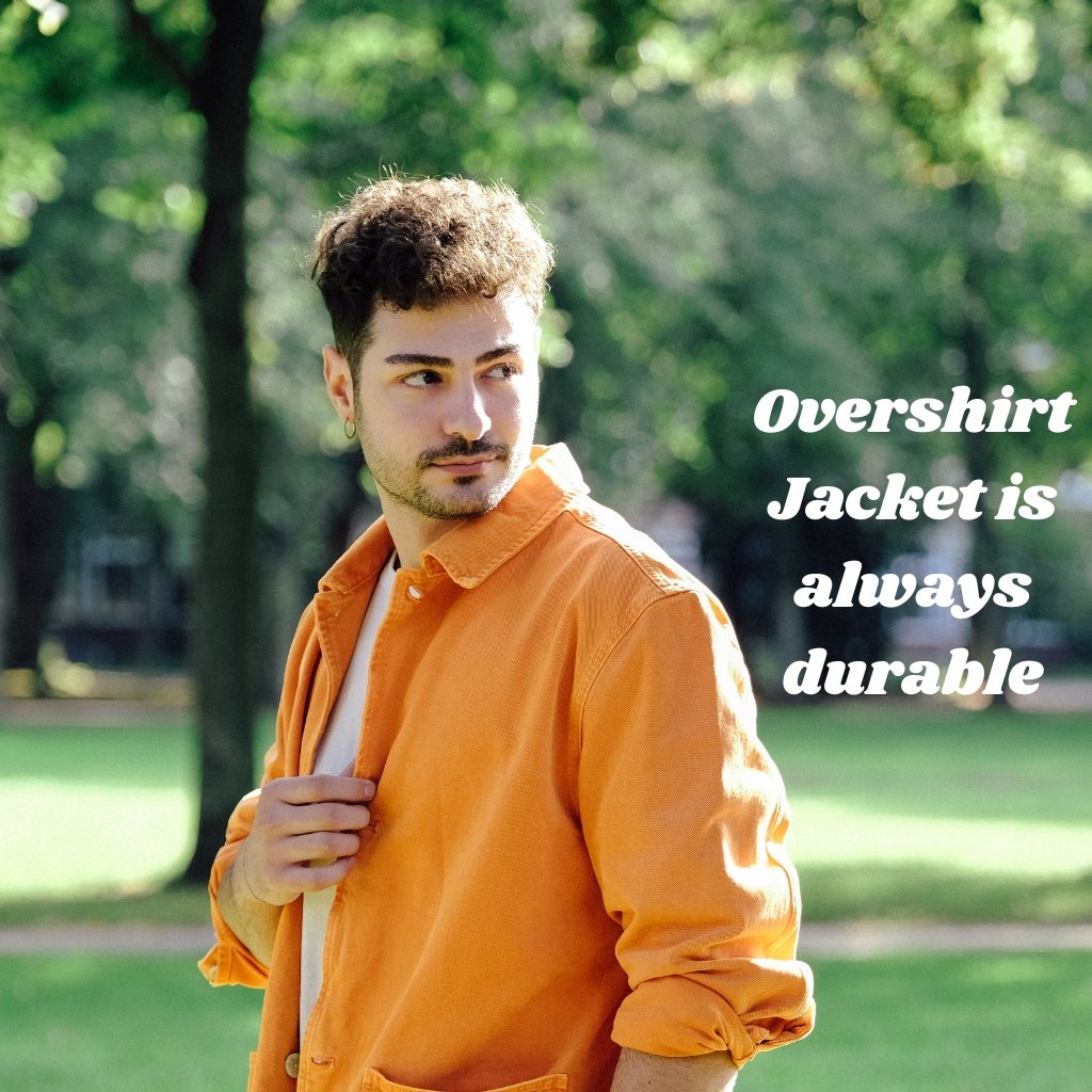A young man stands outdoors in a sunny park, wearing an orange overshirt over a white shirt, looking to the side with a relaxed expression.