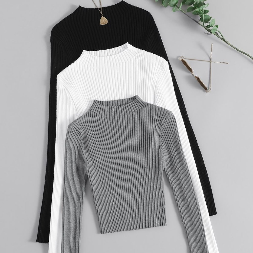 Three ribbed-knit turtleneck sweaters in black, white, and gray stacked together on a flat lay, with a pair of sunglasses and a necklace nearby
