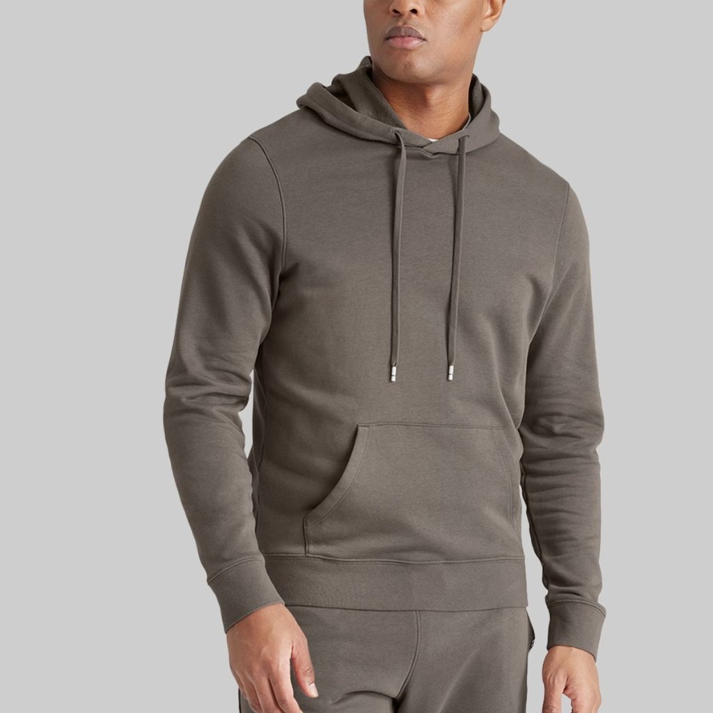 A man wearing a solid charcoal-gray hoodie with a front kangaroo pocket and drawstrings, standing against a light gray background. The outfit appears casual and athleisure-inspired, with matching sweatpants visible at the bottom.