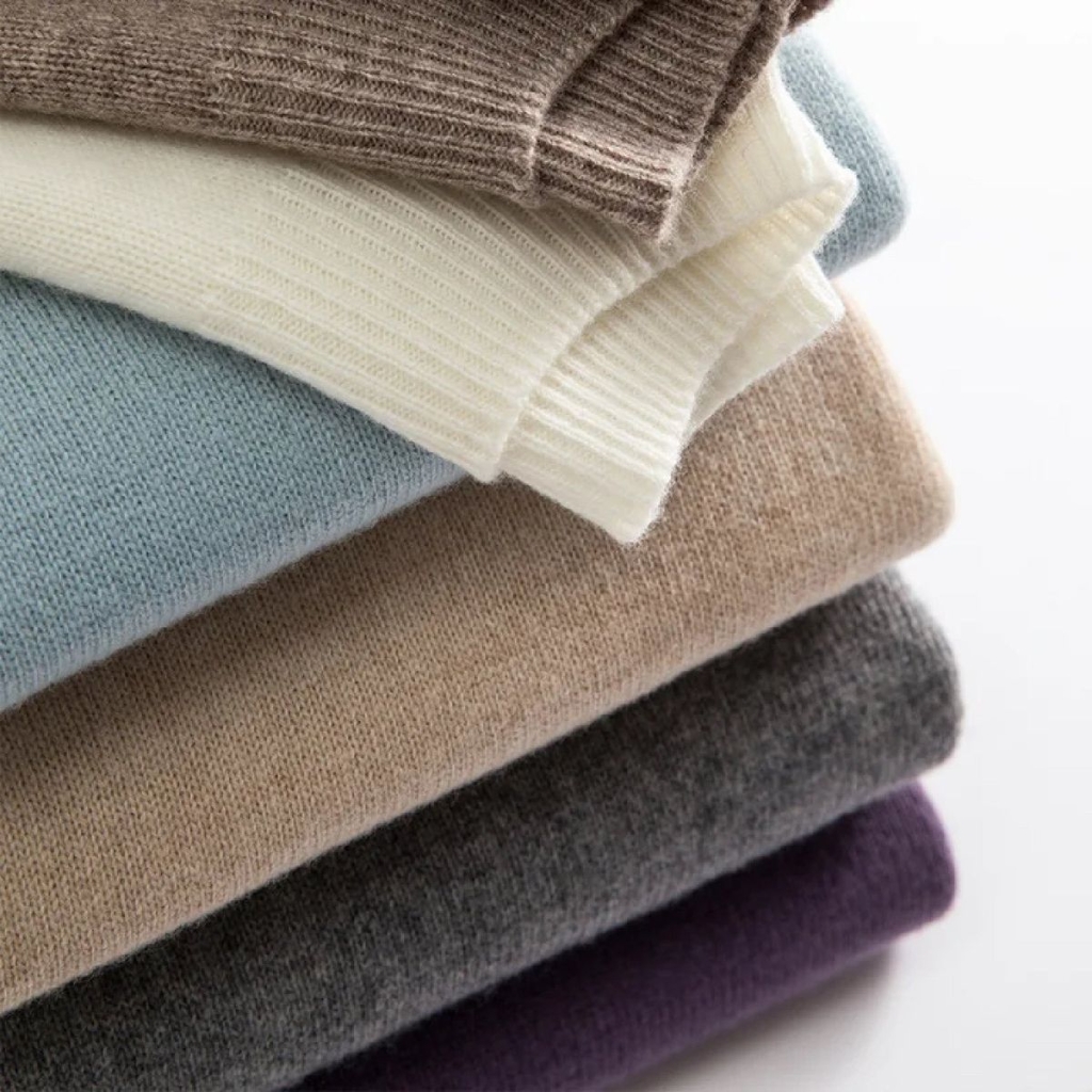 A stack of folded knit sweaters in neutral and muted colors (cream, light blue, beige, gray, black, and plum) photographed from the side