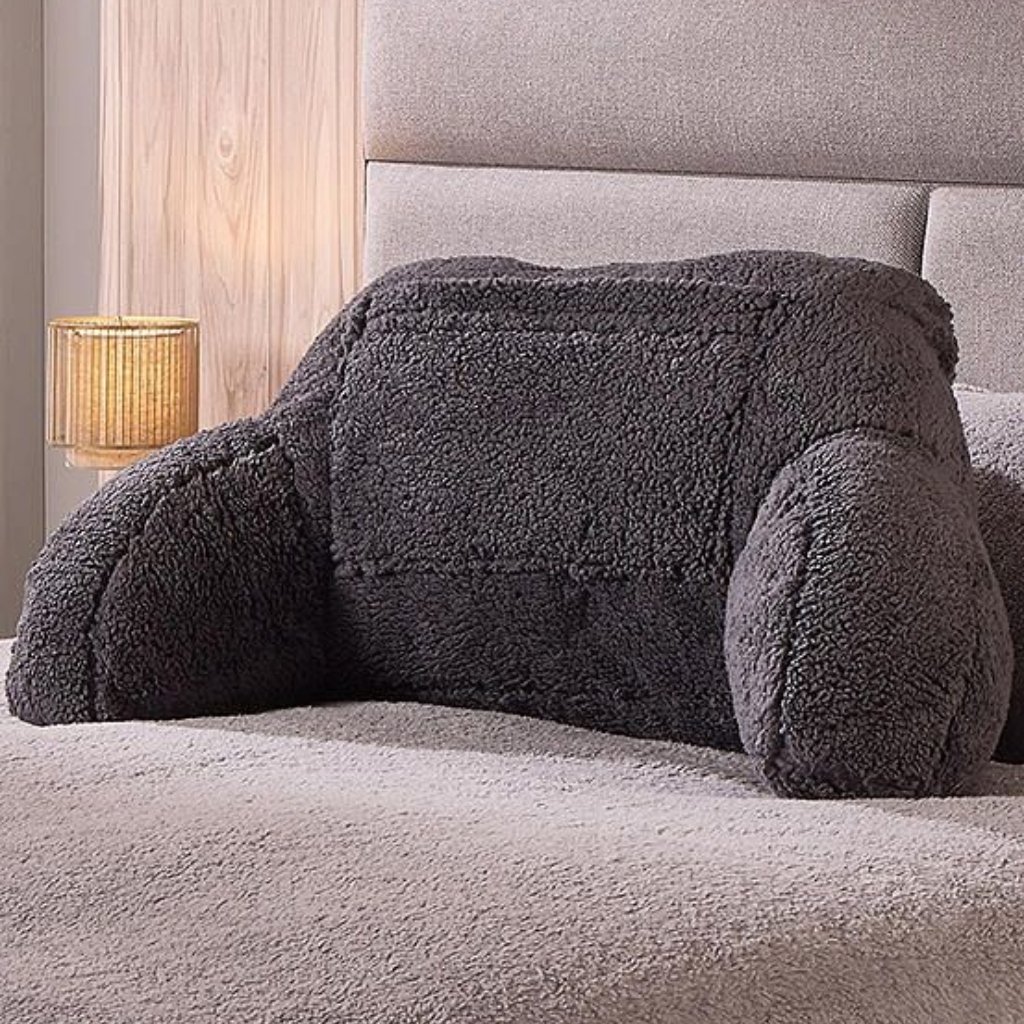 A gray plush stuffed animal shaped like a dog or bear lying face down on a beige bed, with a small lamp on a wooden nightstand in the background.