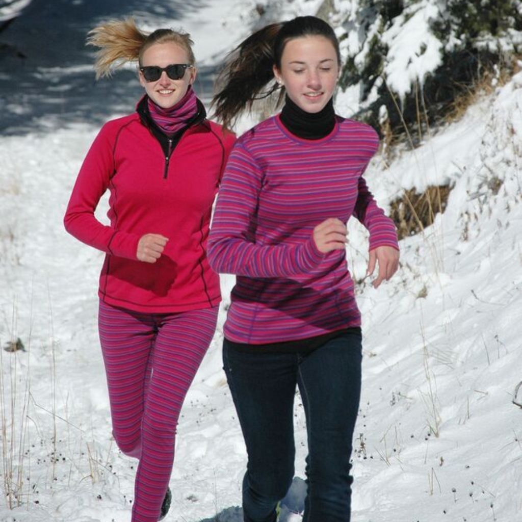 Two women running outdoors in a snowy trail, wearing pink athletic wear; one in a pink long-sleeve top with striped leggings and sunglasses, the other in a striped pink and purple top with dark jeans. Snowy landscape in the background.