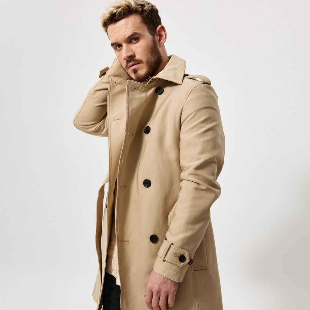  A man wearing a beige trench coat poses sideways against a light background, adjusting the collar with one hand, looking toward the camera.