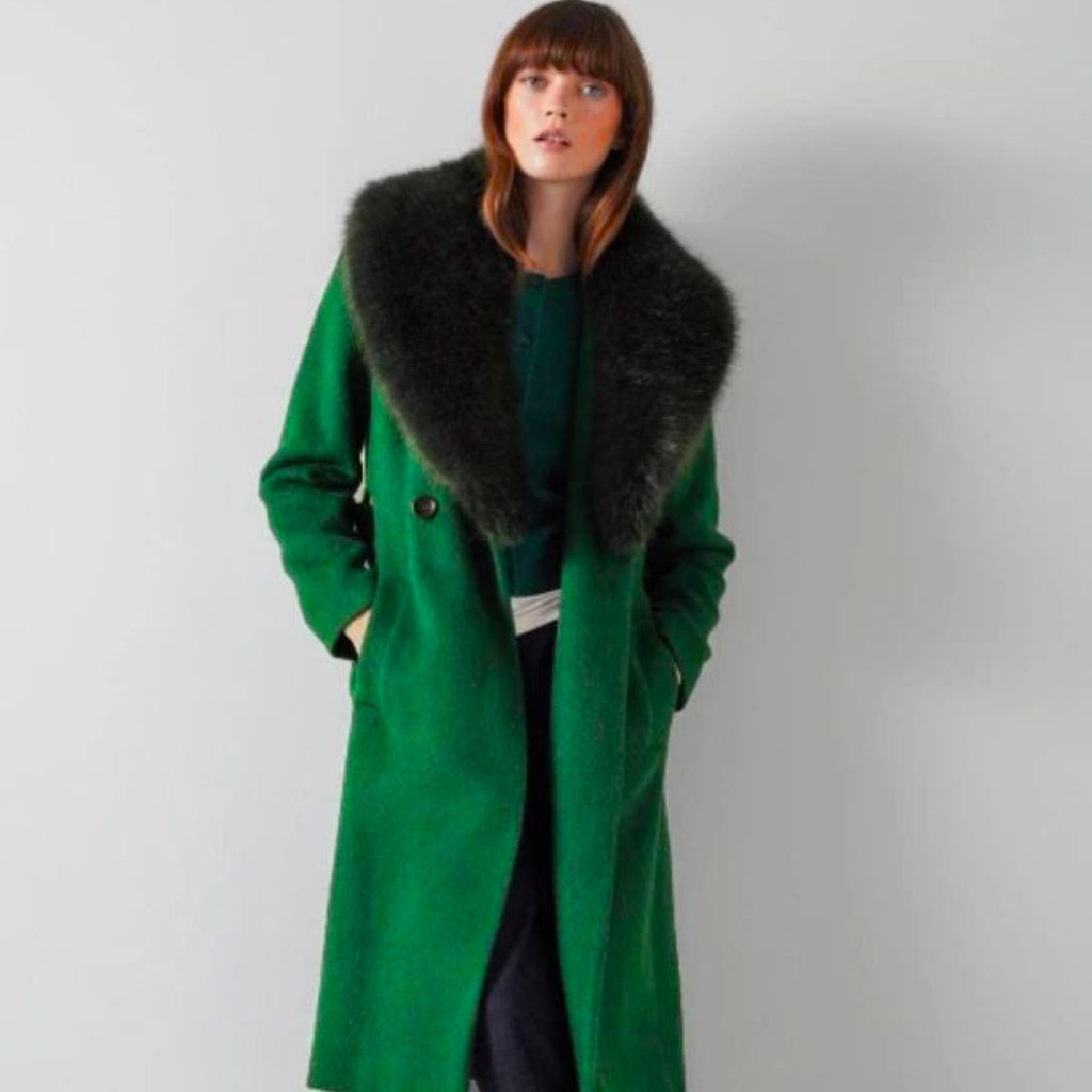 A woman wearing a vibrant green long coat with a large dark fur collar, standing against a light gray wall with hands in her pockets.