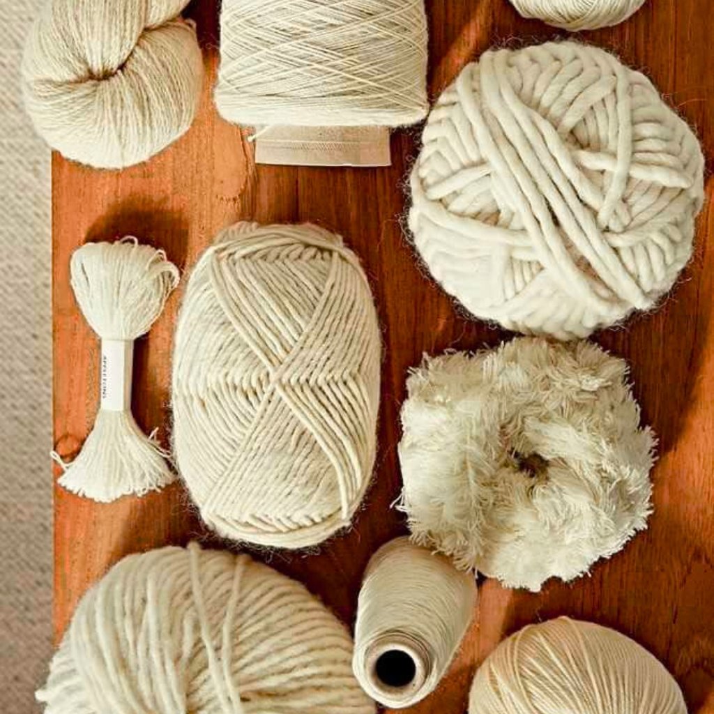 A collection of white or cream-colored yarns and fibers arranged on a wooden surface. Includes skeins, a small tassel, a frayed rope-like piece, a thick loop, and a worn spool, all in natural off-white tones.
