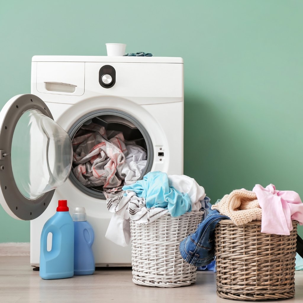 A front-loading washing machine with its door open, two laundry baskets full of clothes spilling over, and detergent bottles placed on the floor nearby against a light green wall.