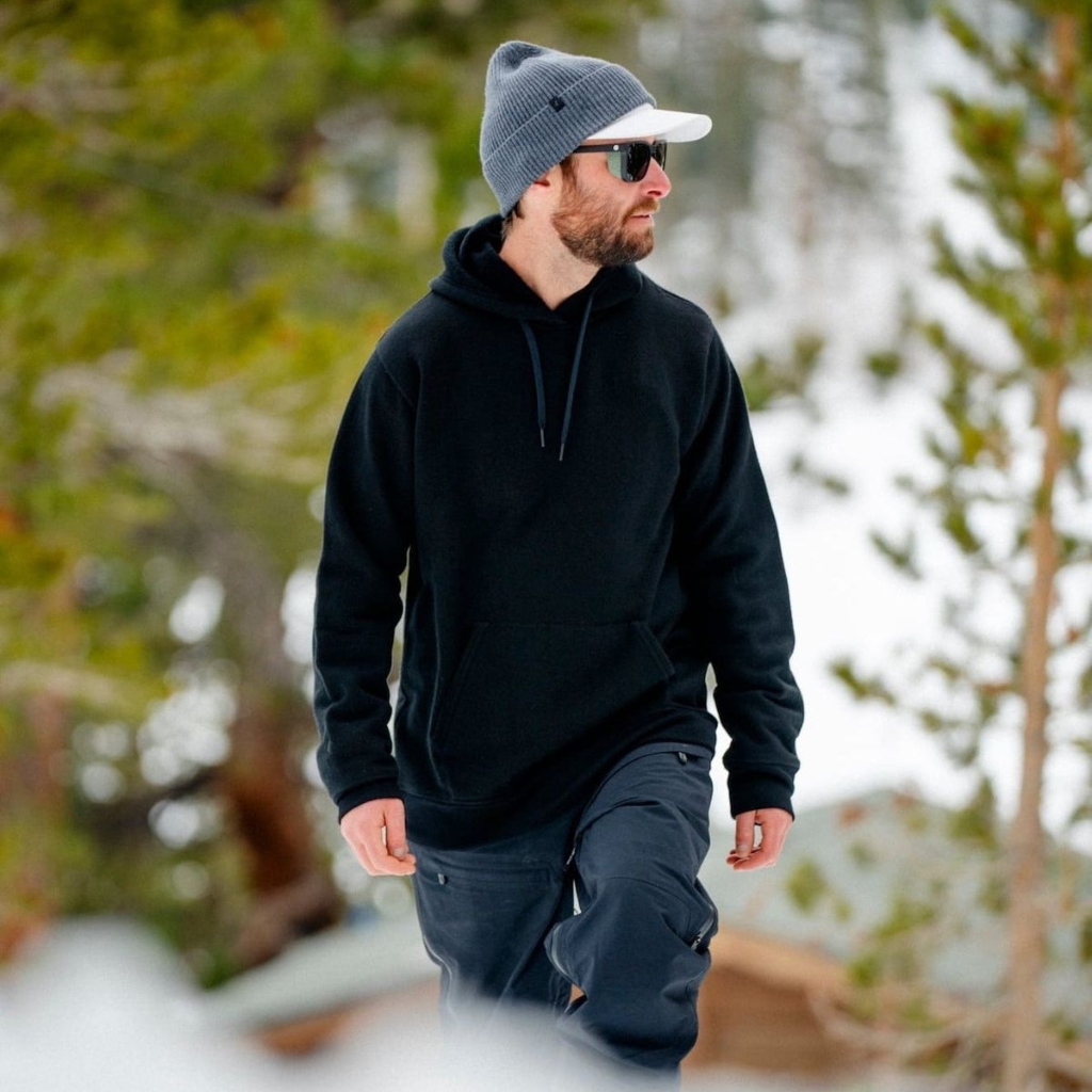 Alt text (for screen readers): A man with a beard wearing a gray knit beanie, dark sunglasses, and a black hoodie, walking outdoors in a snowy, wooded area.

