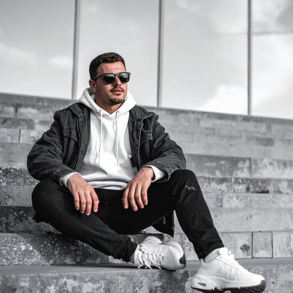  A stylish man sits cross-legged on concrete steps, wearing a white hoodie, black denim jacket, black jeans, white sneakers, and sunglasses.