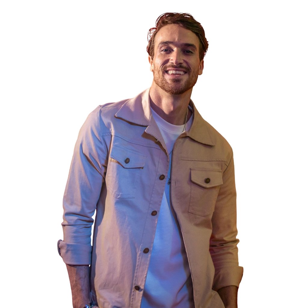 A smiling man standing at a slight angle, wearing a light tan button-up jacket over a white shirt, with rolled sleeves and a relaxed pose.