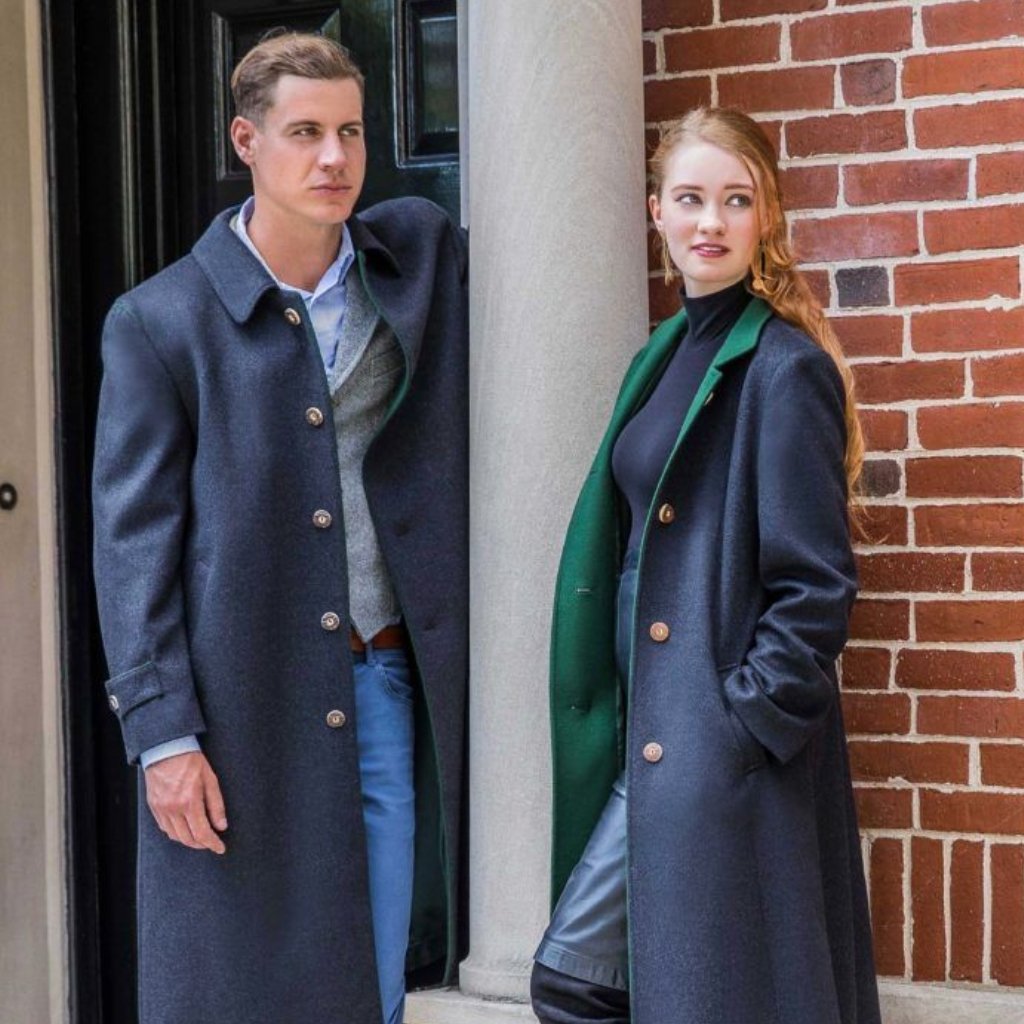 A man and a woman wearing long dark overcoats stand next to a brick wall and a column outside a doorway. The man looks to the left, and the woman looks toward the camera with a slight smile.