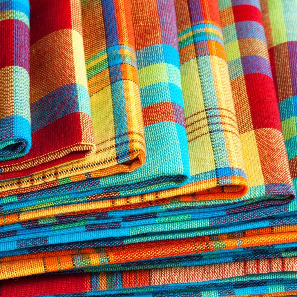 Pile of folded, multicolored plaid fabric with bright blue, orange, red, yellow, and green hues and visible woven texture.