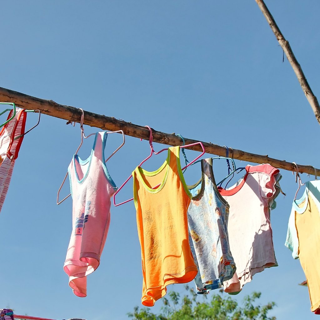 Clothes hang on a clothesline against a clear blue sky, including pink, orange, and light-colored tank tops and shirts with colorful hangers.