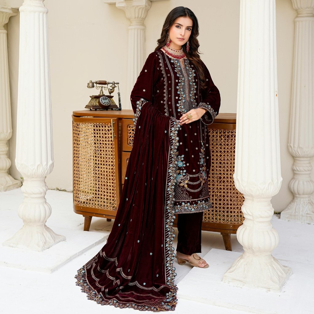 A woman wearing a burgundy traditional embroidered ensemble with a matching long shawl, standing beside a vintage wooden cabinet and cream columns.