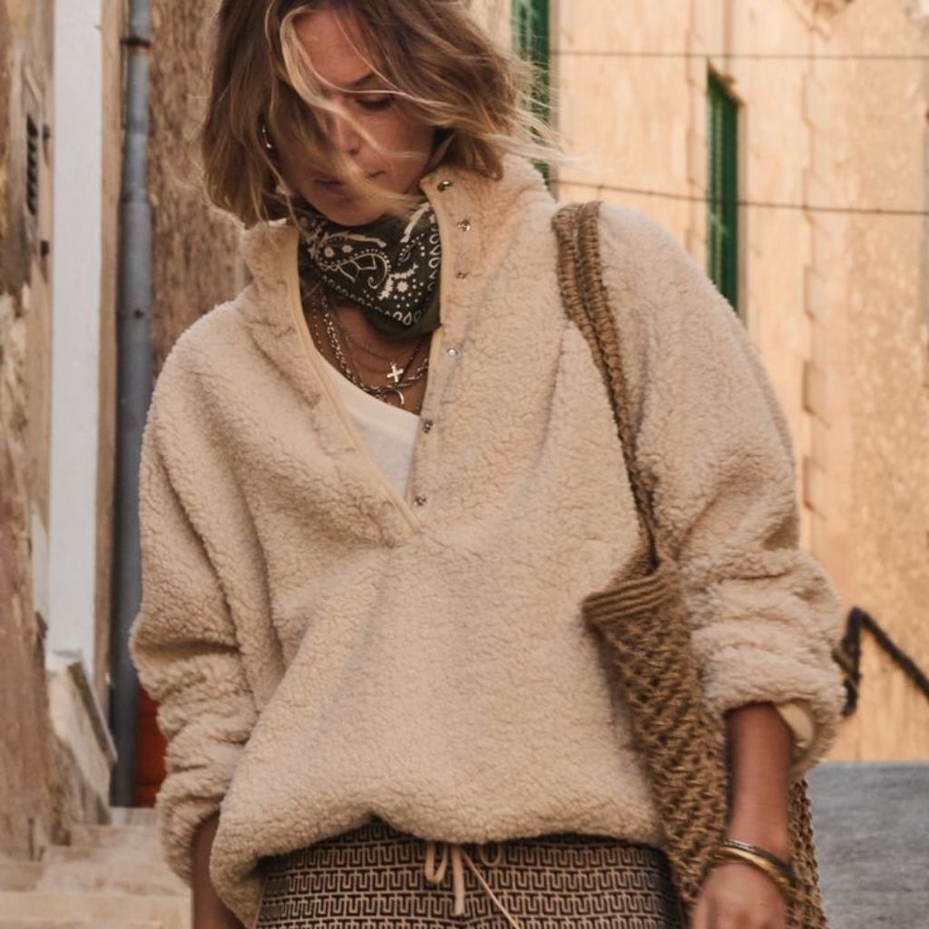  A stylish person walking down a sunlit street wearing a cream fuzzy pullover with a V-neck and white top underneath. They have a green bandana around the neck, layered necklaces, and a woven tote bag slung over one shoulder. The individual wears patterned drawstring shorts or loungewear below and has tousled blond hair partially obscuring their face.